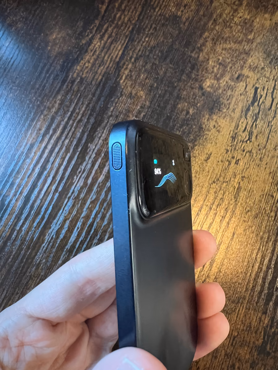 The Comu Action Pro seen from the side in a hand, its screen showing a battery level and a waveform.