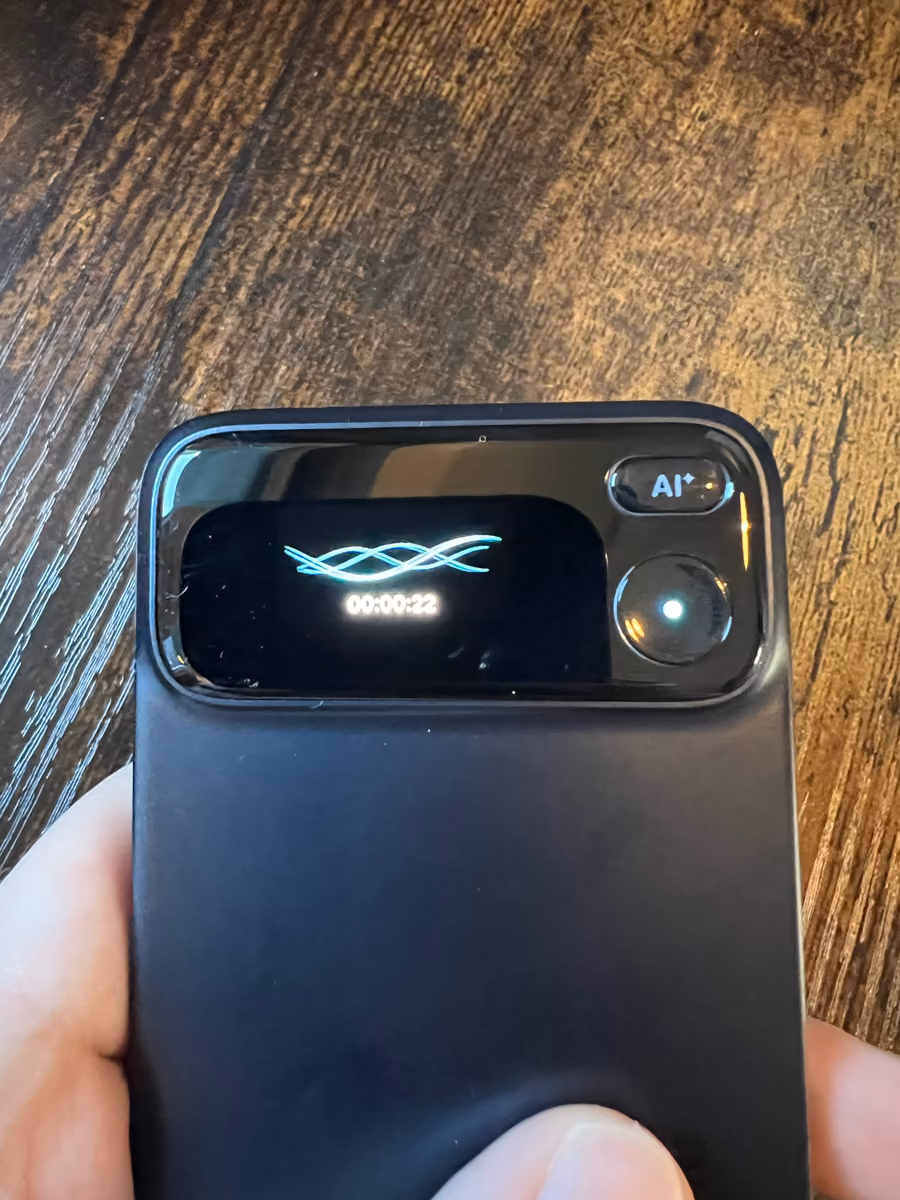 The Comu Action Pro held in a hand, its OLED screen showing a recording timer and the AI button.